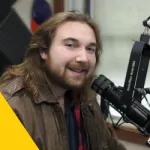 A man with long brown hair and a beard, wearing a brown jacket, smiles while sitting at a radio station microphone. Acoustic foam panels and a window are visible in the background. A yellow triangle appears in the bottom left corner.