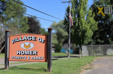 An orange sign reads Village of Homer Streets & Parks beside a road, with trees, a flagpole, and a fence in the background. A radio station logo X101 Always Classic is in the top right corner.