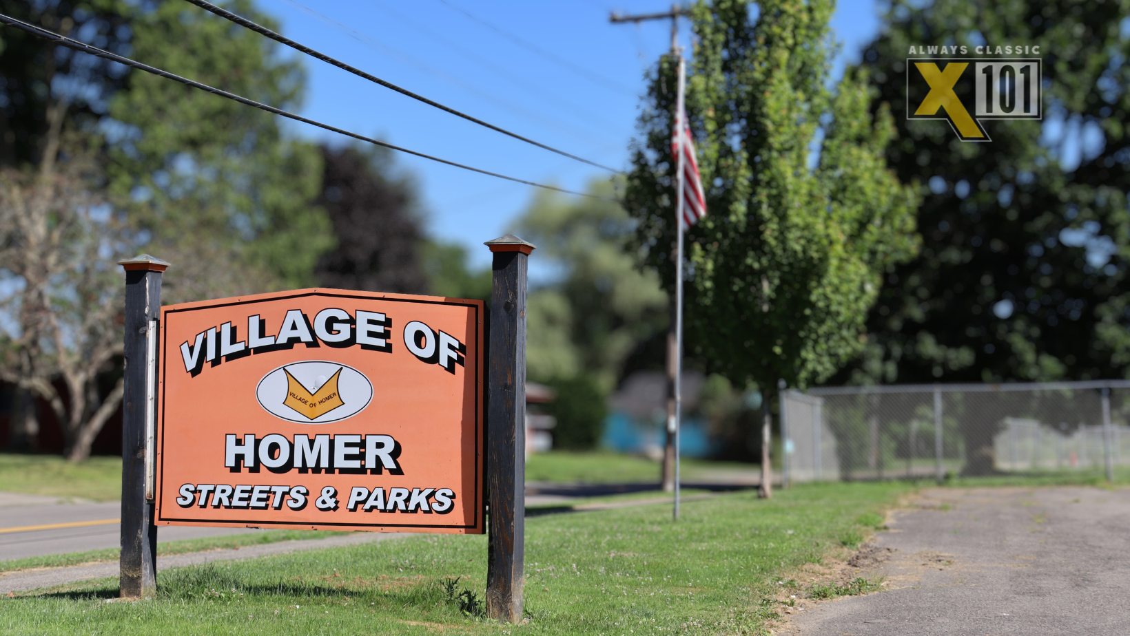 Amnesty Day For Village of Homer Residents Coming up in October 7 An orange sign reads Village of Homer Streets & Parks beside a road, with trees, a flagpole, and a fence in the background. A radio station logo X101 Always Classic is in the top right corner.